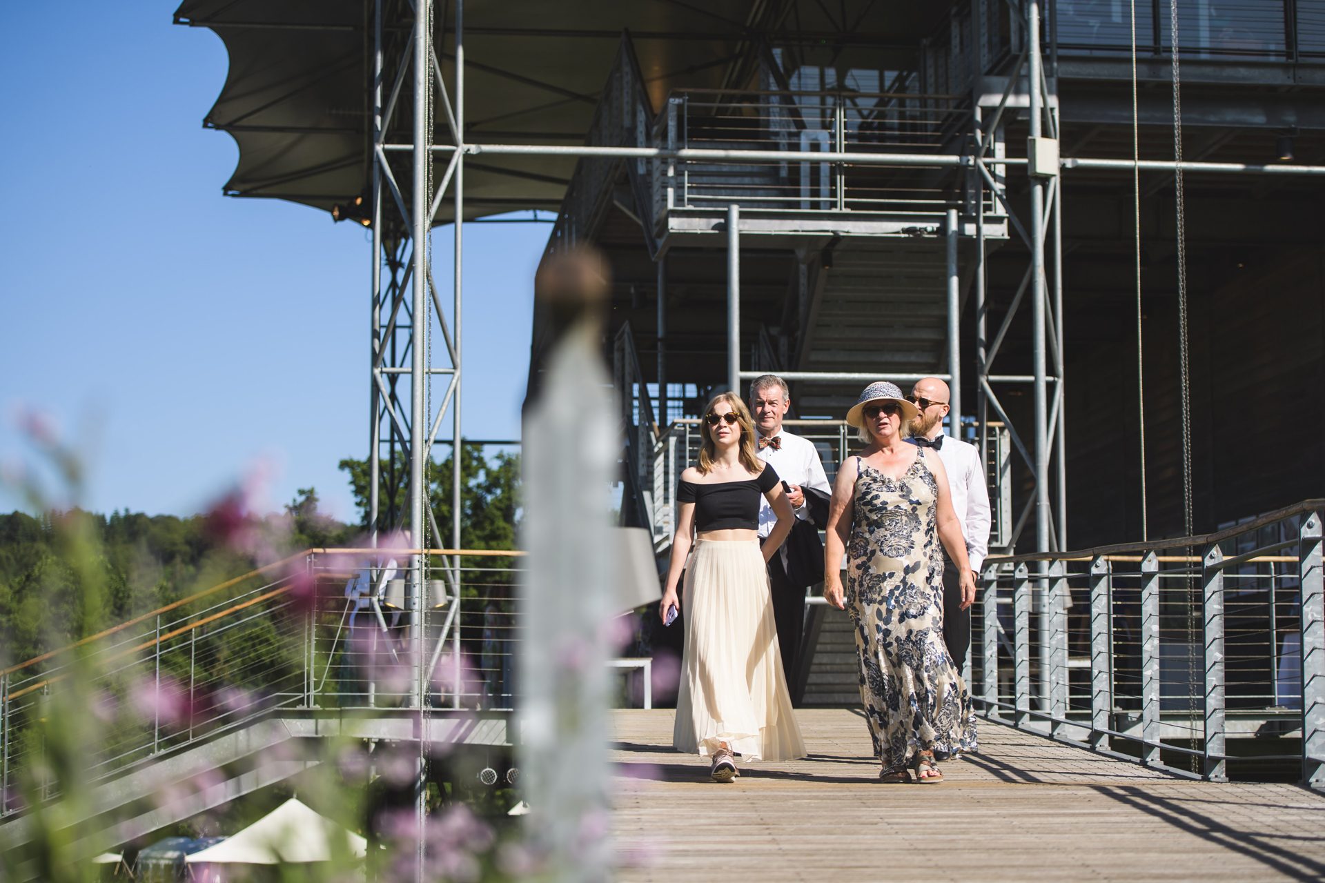 Four people walking over a wooden bridge with blue sky and the opera house in the background