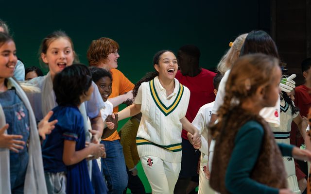 Girl wearing cricket sweater and white trousers running towards the front of the stage whilst smiling and surrounded by cheering friends.