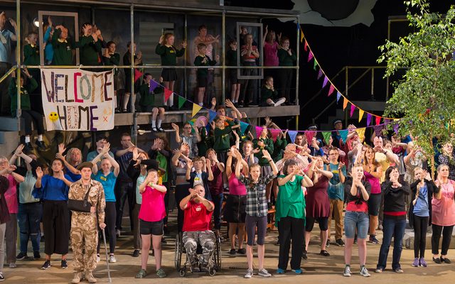 A large group of adults and young people stand together on stage. To the right there is a lush green tree and to the left a scaffold with a banner reading 'Welcome Home'. An image from Garsington Opera's 2017 production of 'Silver Birch' by Panufnik.