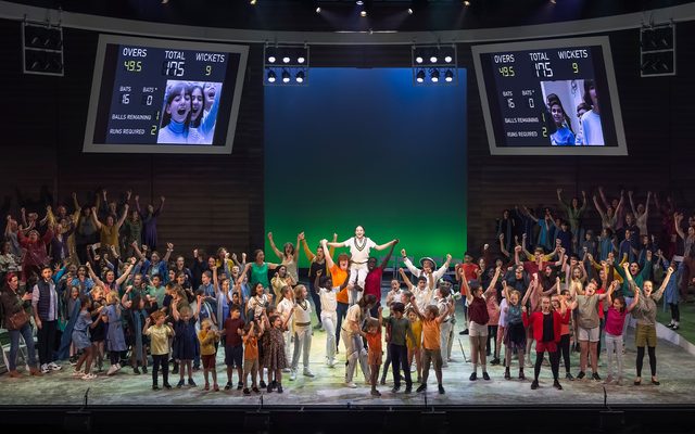 The whole stage is full of people with their arms raised in the air as if cheering. In the centre a young girl dressed in white cricket scrubs is lifted by a person either side above the crowds around them. An image from Garsington Opera's 2022 production of 'Dalia' by Panufnik.