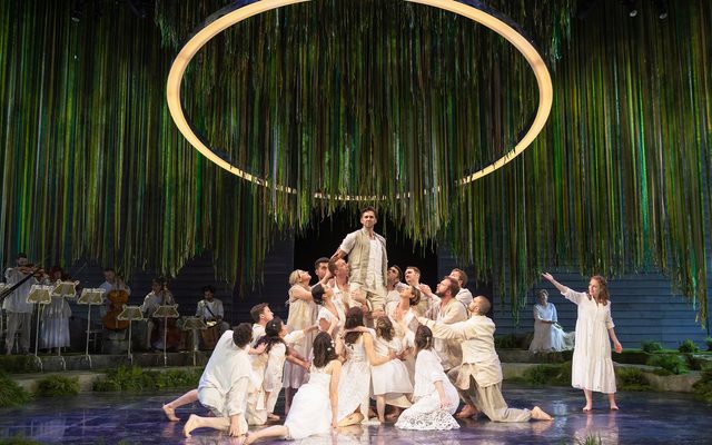 Under a large, glowing gold circle, a group of people in white flowing clothes gather in a pyramid, looking up to the man stood at it's top. An image from Garsington Opera's 2022 production of 'Orfeo' by Monteverdi.