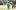 GO35-3 A group of seven young people walk towards the camera through the beautiful, green Wormsley grounds with the opera pavilion standing to their right in the background and just out of focus. They walk together in pairs in a loose line, fading out of focus towards the back. All are dressed in elegant evening wearing and carrying full champagne glasses.