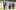 A group of seven young people walk towards the camera through the beautiful, green Wormsley grounds. They walk together in pairs in a loose line, fading out of focus towards the back. All are dressed in elegant evening wearing and carrying full champagne glasses.