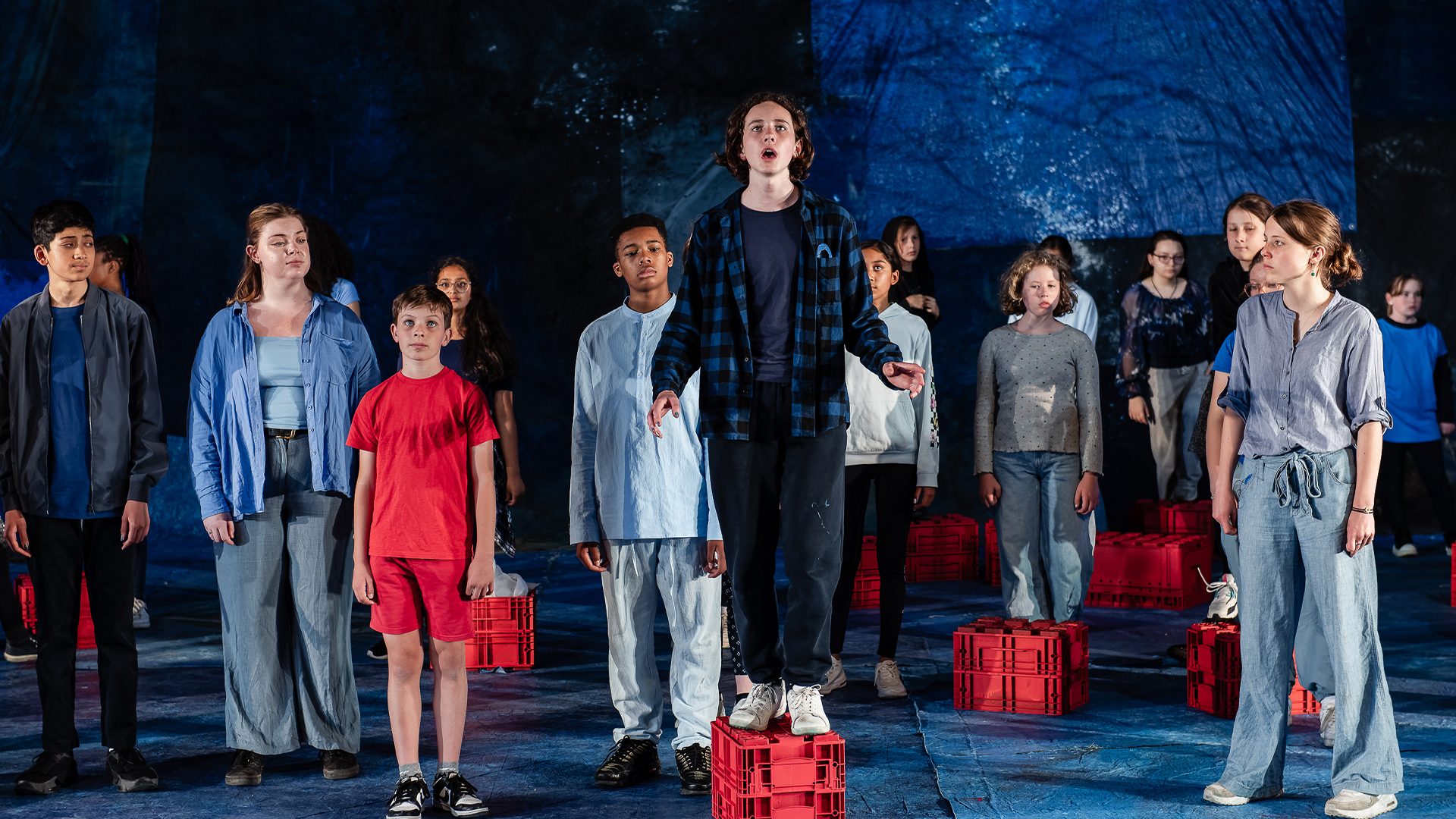 A group of young people, dressed in blue, stand on a stage covered in old tarpaulins and red crates. In teh centre a boy in a blue plaid shirt and with long brown hair stands on a red crate singing. To the left of him stands a young boy dressed all in red, staring in front of himself. An image from Garsington Opera's 2023 Youth Company performance of 'The Moon is Listening' by Richard Taylor.