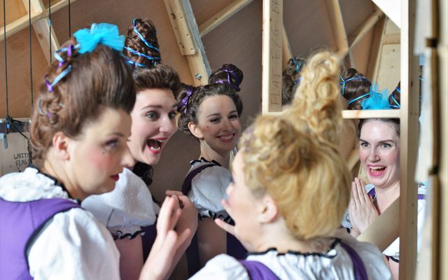 Five women with their hair up in two domes on their heads, laugh together in a backstage area.