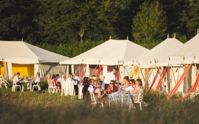 A view down the dining tents at Wormsley. Golden sun lights the white tents and people picnic both inside and outside them.