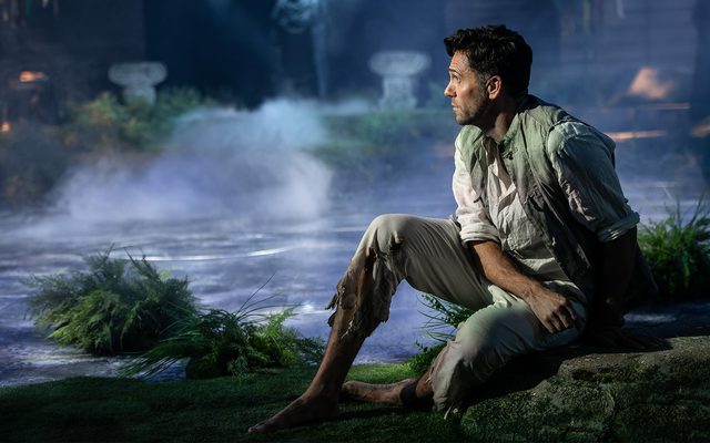 A man crouches on a bank by the edge of a dark lake. An image from Garsington Opera's 2022 production of 'Orfeo' by Monteverdi.
