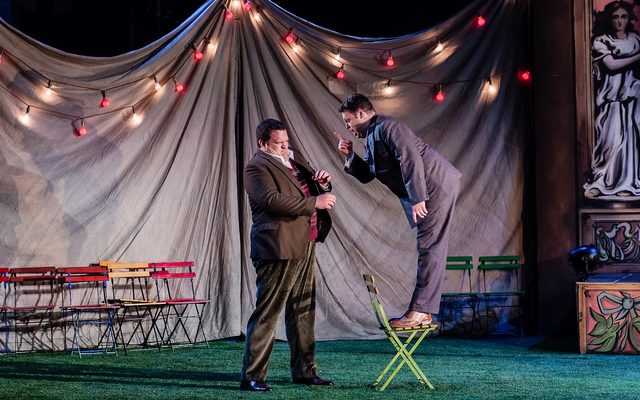 A man in a dark grey suit stands on a chair to berate a taller man, who shies away from his outstretched finger. An image from Garsington Opera's 2023 performance of 'The Bartered Bride' by Smetana.