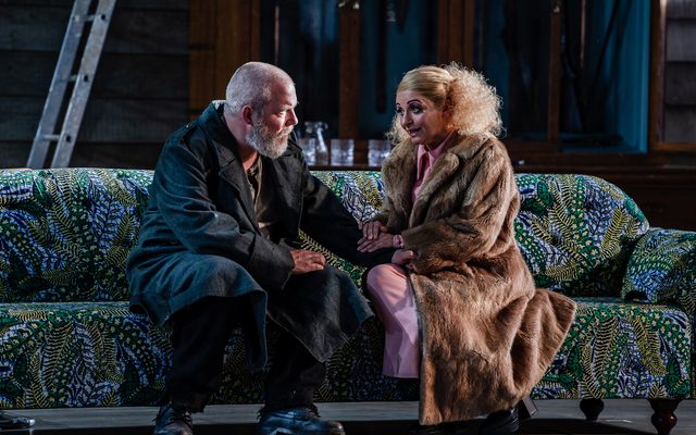 A man in a trench coat and a woman in a brown fur coat sit together on a lavish green sofa, comforting one another. An image from Garsington Opera's 2023 performance of 'Mitridate, re di Ponto' by Mozart.