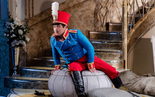 A man in a bright blue military jacket, with vivid red trousers and hat sits leaning forward on a poof. An image from Garsington Opera's 2023 performance of 'Il barbiere di Siviglia' by Rossini.