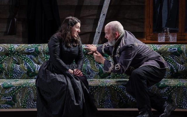 A woman in a long black dress sits next to a man in military dress on a maximalist green sofa. An image from Garsington Opera's 2023 performance of 'Mitridate, re di Ponto' by Mozart.