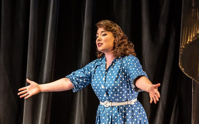 Lady with brown curly hair in a blue dress with white spots on it gesturing towards the audience in The Bartered Bride