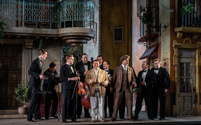 Two gentleman dressed in light brown suits stand in the middle of a group of people dressed as musicians and holding instruments. An image from Garsington Opera's 2023 performance of 'Il barbiere di Siviglia' by Rossini.
