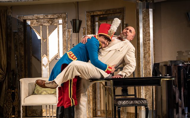 A man dressed in a bright blue military jacket with a big red hat, hugs a very surprised looking gentleman dressed in a cream suit. An image from Garsington Opera's 2023 performance of 'Il barbiere di Siviglia' by Rossini.