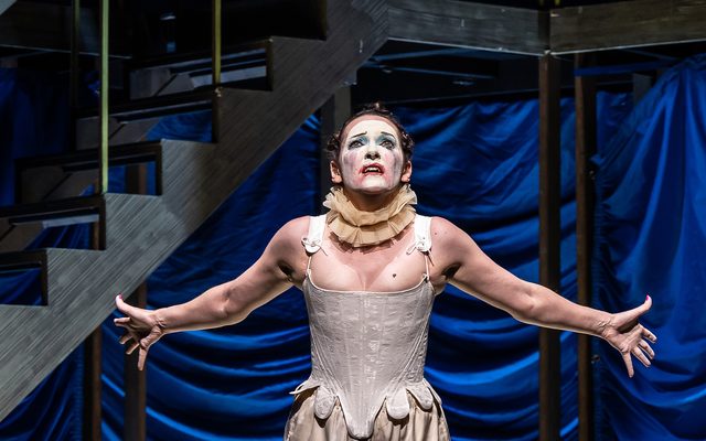 A woman in a white corset with smudged clown makeup throws her arms wide. An image from Garsington Opera's 2023 performance of 'Ariadne auf Naxos' by Strauss.