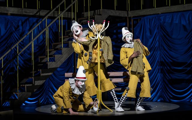 Three people dressed in yellow clown outfits with white painted faces mess around with some discarded clothes and a coat stand. An image from Garsington Opera's 2023 performance of 'Ariadne auf Naxos' by Strauss.