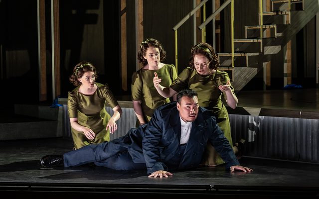 Three figures dressed in green, fitted dress gather around a man in a dark blue suit, prostrated on the ground. An image from Garsington Opera's 2023 performance of 'Ariadne auf Naxos' by Strauss.