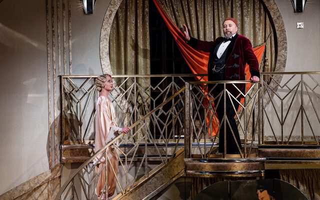 A man in a deep red velvet smoking jacket stands dramatically at the top of a staircase as if ready to sing. An image from Garsington Opera's 2023 performance of 'Il barbiere di Siviglia' by Rossini.