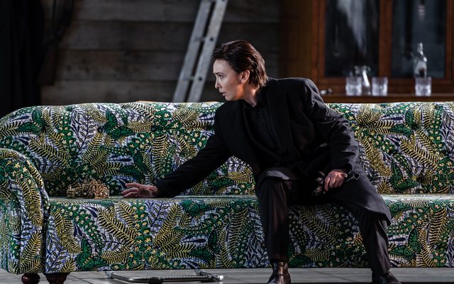 A figure dressed in black sits on a highly patterned green sofa, stretching out a hand for the crown next to them. An image from Garsington Opera's 2023 performance of 'Mitridate, re di Ponto' by Mozart.