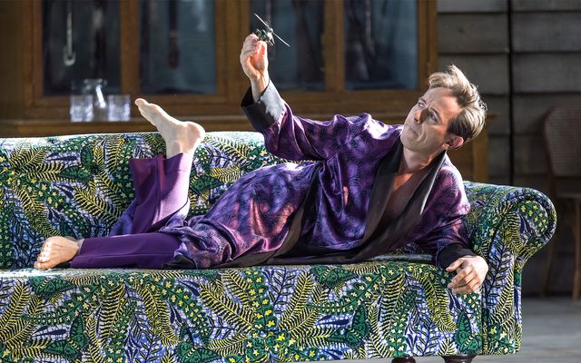 A man in a long, purple silt dressing gown lies on a vivid green sofa playing with a toy helicopter. An image from Garsington Opera's 2023 performance of 'Mitridate, re di Ponto' by Mozart.