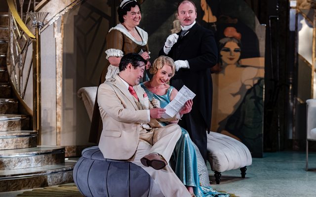 A couple in 1920s evening wear sit on a poof reading a document. Behind them stand to people dressed as a maid and a butler. An image from Garsington Opera's 2023 performance of 'Il barbiere di Siviglia' by Rossini.