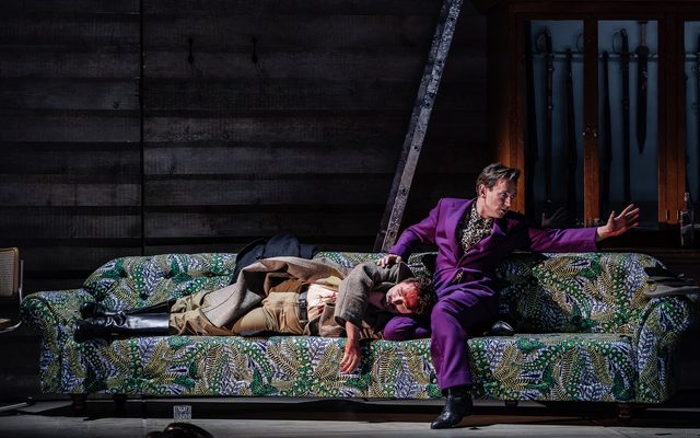 A man in a purple suit sits on a bright green sofa and stretches out a hand to the right. Next to him is the body of a man in beige military uniform, his forehead covered in blood. An image from Garsington Opera's 2023 performance of 'Mitridate, re di Ponto' by Mozart.