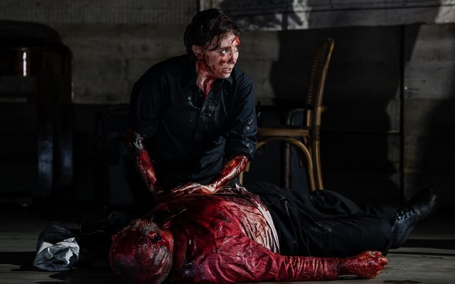 A figure dressed in black crouches over the body of a man. Both are covered in blood. An image from Garsington Opera's 2023 performance of 'Mitridate, re di Ponto' by Mozart.