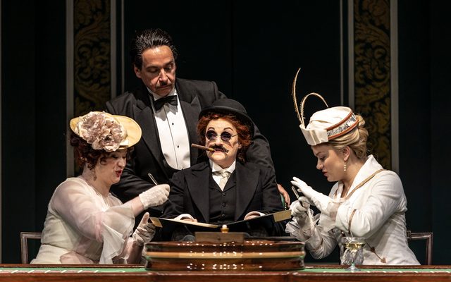 A scene from Così fan tutte in 2022, this image shows a woman dressed up as a male lawyer tricking two women into signing a contract with a man peering over.