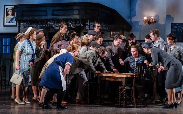 A man sits at a pub table, surrounded by a large crowd of people. An image from Garsington Opera's 2023 performance of 'The Bartered Bride' by Smetana.