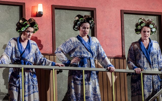 Three women in white and blue silk dressing gowns with giant hair-curlers in stand along a mezzanine balcony, staring down. An image from Garsington Opera's 2023 performance of 'Ariadne auf Naxos' by Strauss.