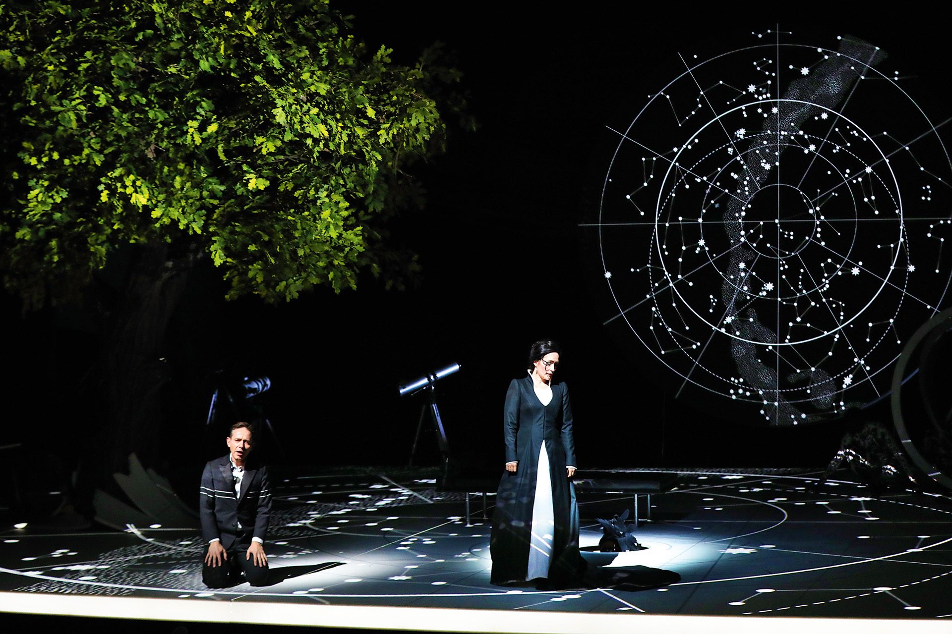 A woman in a black and white dress stands in the centre of a dark, sparse stage, while a man kneels underneath a vivd green tree. Both are surrounded by constellations.