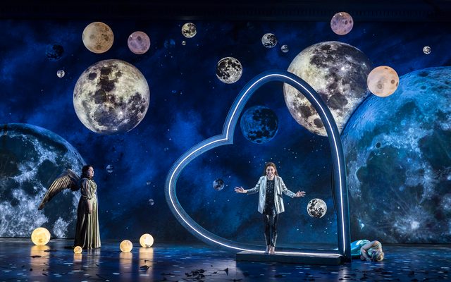 A woman in a silver jacket and black trousers standing in a giant heart surrounded by planets in the sky, on stage in Semele.