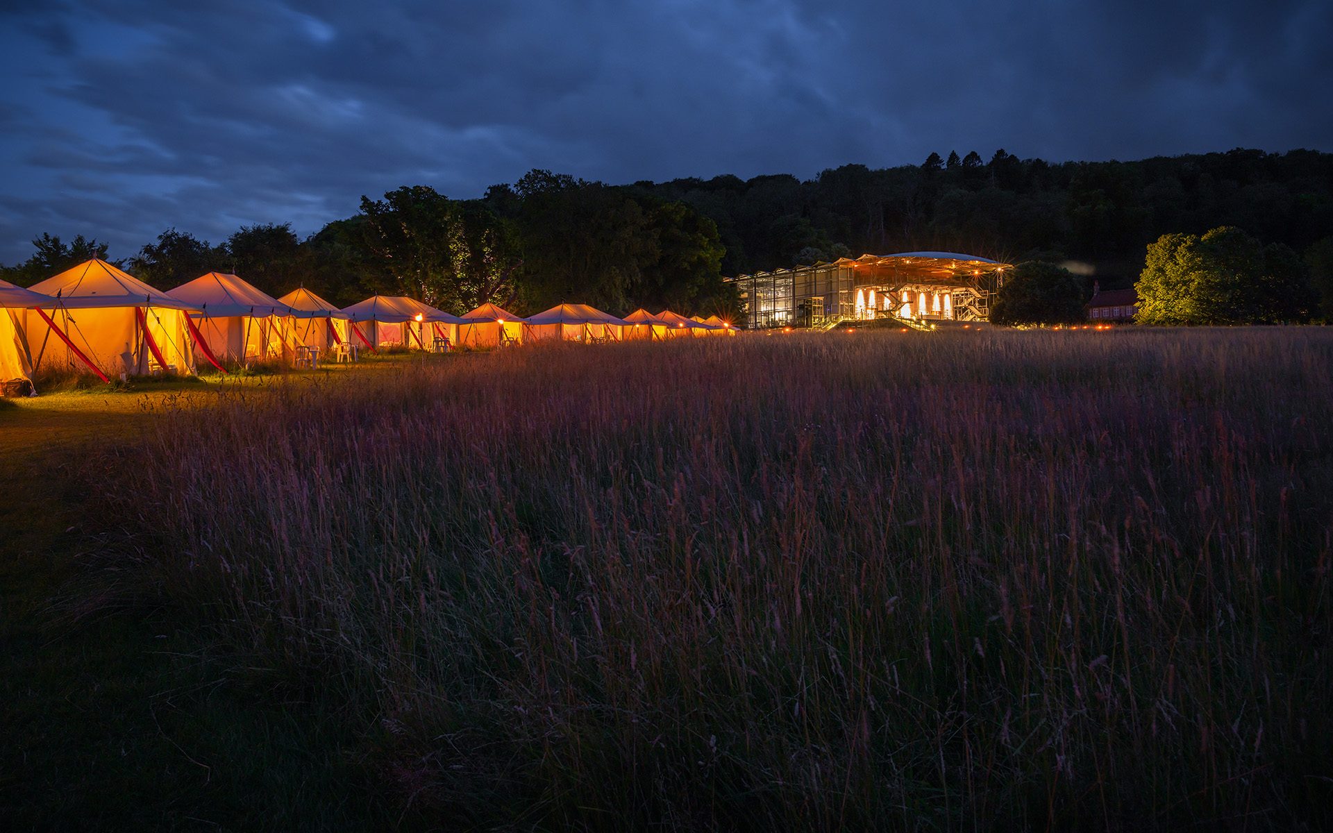 A line of warmly lit tents snake through the darkness towards the brightly lit opera pavilion in the distance.