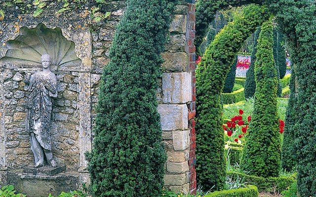 A stone statue in an alcove is backgrounded by a lush, green garden, at Garsington Manor.