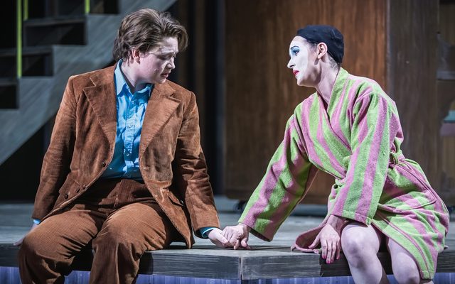 Two people sit on the edge of the stage, one dressed in a brown cord suit with a blue shirt, the other in a green and pink striped dressing gown with a painted, white face. An image from Garsington Opera's 2023 performance of 'Ariadne auf Naxos' by Strauss.