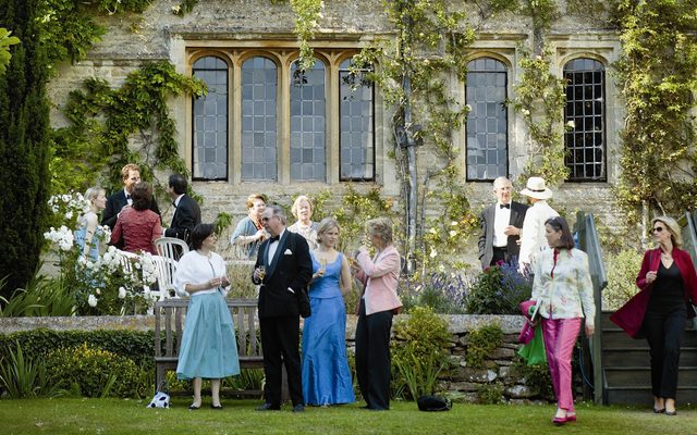 People dressed in elegant evening wear dress gather, drinking champagne, outside the beautiful Garsington Manor.