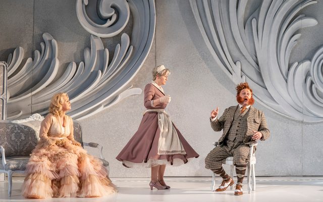 From Der Rosenkavalier, a blonde lady sitting on an opulent sofa with a big skirt, with a tweed wearing ginger man sitting on the chair opposite, and a maid dressed in pink in between, looking outraged at what the man is saying.