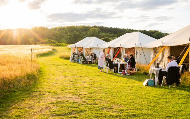 Golden late evening sun streams down on to some of the dinning tents at Wormsley while people picnic both within and without them.