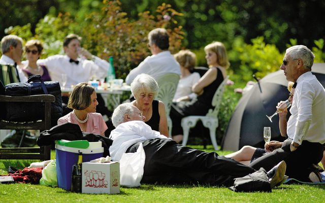People dressed in evening dress lounge on lush green grass, having a picnic at Garsington Manor.