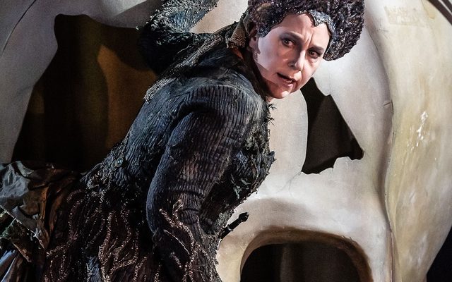 A woman dressed in dark clothes with a pointed headdress rest a hand on a giant skull as she passes. This is Ježibaba, the witch, played by Christine Rice in our 2022 Rusalka.