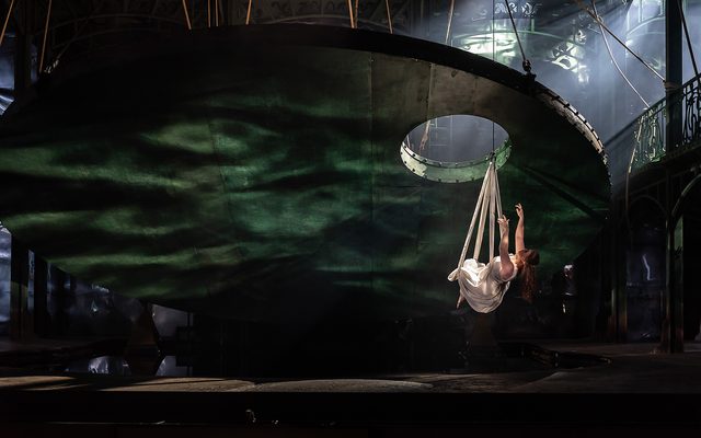 A woman in white is lowered on white ribbons through a hole in a giant suspended disk towards a pool of water.