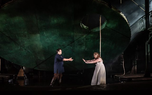 A woman in a white dress reaches for a man dressed in blue, both stood within a green pool onstage.