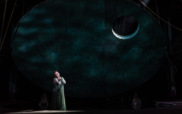 A woman dressed in a green dress sings up to the moon above her.