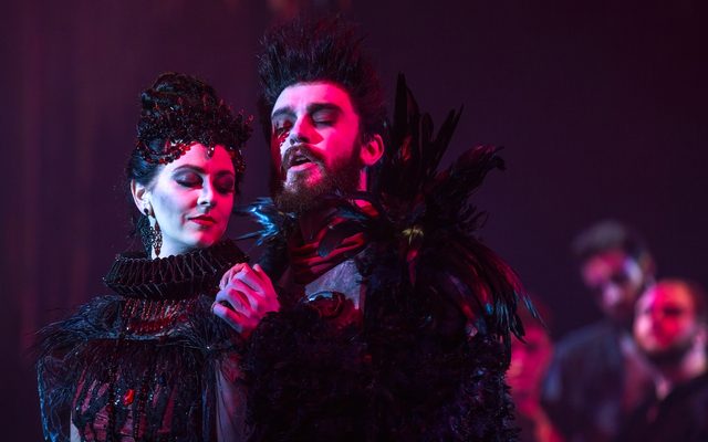 A man embraces a woman from behind, both dressed in black and lit by an ethereal pink light.
