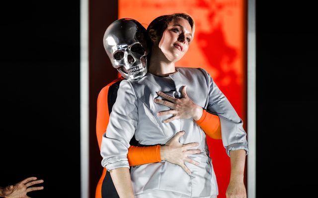 A figure in a silver skull mask grabs a woman dressed in silver from behind against an orange backdrop.