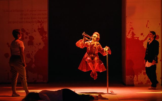 A figure dressed as a court herald in orange hovers unsuspended in the middle of the stage. He blows a trumpet.