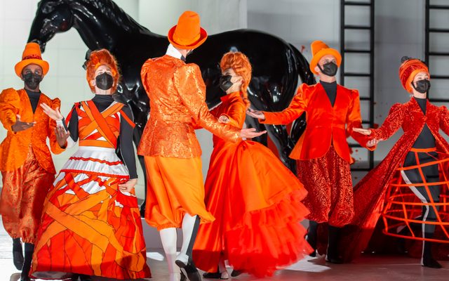 Men and women dressed in vivid orange period costumes dance against a black and white set.